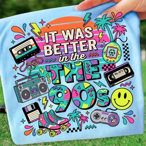 May include: A light blue t-shirt featuring a colorful graphic with the text "IT WAS BETTER in the 90s". The design includes illustrations of a cassette tape, a boombox, a floppy disk, roller skates, a handheld game, a VHS tape, and a smiley face.