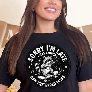 May include: Black t-shirt with a circular graphic featuring a raccoon on a skateboard holding a mug. The graphic includes the text "SORRY I'M LATE I WAS AVOIDING NON-PREFERRED TASKS" in a white, stylized font. The raccoon is depicted in shades of gray and white.