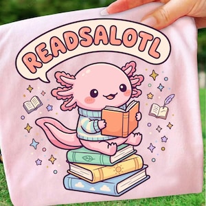 May include: Pink t-shirt featuring a cartoon axolotl reading a book, sitting on a stack of books. The axolotl wears a blue and white striped sweater. The text "READSALOTL" is in a speech bubble above the axolotl.