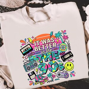 May include: A white crewneck sweatshirt featuring a colorful graphic print with the text "IT WAS BETTER in the 90s". The design includes illustrations of a cassette tape, boombox, floppy disk, roller skates, handheld game, and a yellow smiley face.