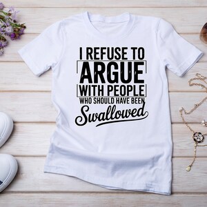 Può includere: T-shirt bianca con la scritta nera "I REFUSE TO ARGUE WITH PEOPLE WHO SHOULD HAVE BEEN SWALLOWED". La maglietta è esposta su una superficie di legno chiaro con altri accessori.