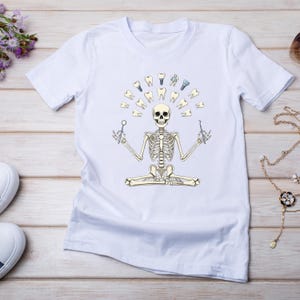 May include: White t-shirt with a humorous dental design. It features a skeleton in a yoga pose, encircled by teeth and dental tools. The skeleton is holding dental instruments. A fun take on anatomy.