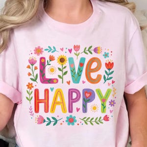 K&ouml;nnte beinhalten: Hellrosa T-Shirt mit den Worten "LIVE HAPPY" in bunten, floral dekorierten Buchstaben. Das Design umfasst Blumen, Herzen und Bl&auml;tter und erzeugt eine fr&ouml;hliche und lebendige &Auml;sthetik. Das Shirt hat kurze &Auml;rmel.