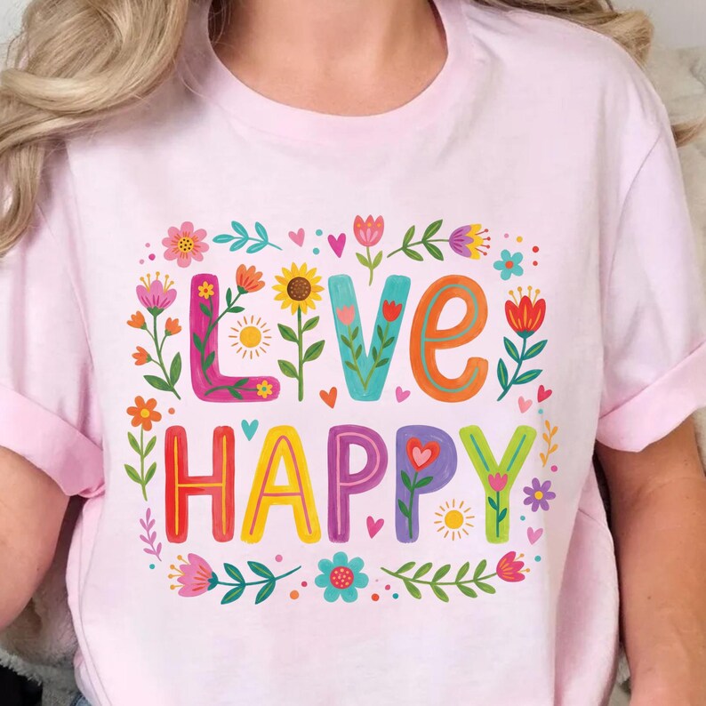 K&ouml;nnte beinhalten: Hellrosa T-Shirt mit den Worten "LIVE HAPPY" in bunten, floral dekorierten Buchstaben. Das Design umfasst Blumen, Herzen und Bl&auml;tter und erzeugt eine fr&ouml;hliche und lebendige &Auml;sthetik. Das Shirt hat kurze &Auml;rmel.