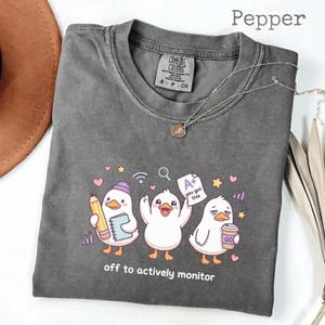 May include: Pepper-colored t-shirt with a graphic of three cartoon ducks, each with a different accessory. One duck holds a pencil and notebook, another holds an A+ paper, and the last holds a coffee cup. The text "off to actively monitor" is printed below the ducks.