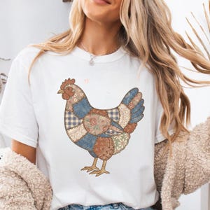 May include: White t-shirt with a patchwork chicken design. The chicken is made up of various fabric patterns in blue, brown, and floral prints. It has a red comb and yellow feet, giving a rustic, country style appearance.