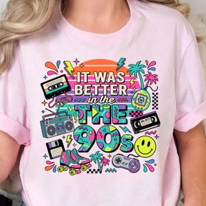May include: A light pink t-shirt featuring a colorful graphic print with the text IT WAS BETTER in the 90s. The design includes a cassette tape, boombox, floppy disk, roller skates, handheld game, VHS tape, smiley face, and game controller.