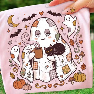 May include: A light pink t-shirt featuring a graphic of a ghost wrapped in a patchwork quilt, holding a steaming mug of coffee with a black cat resting on its lap. The design includes small ghosts, bats, pumpkins, mushrooms, and stars in autumn colors.
