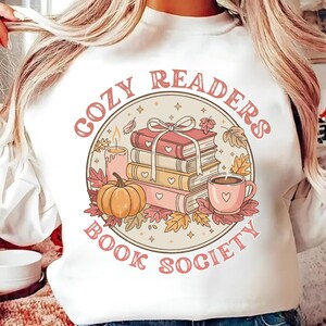 May include: White sweatshirt with a circular graphic reading "Cozy Readers Book Society." The design includes a stack of books, a pumpkin, a coffee cup, a candle, and autumn leaves in pink, orange, and brown hues.