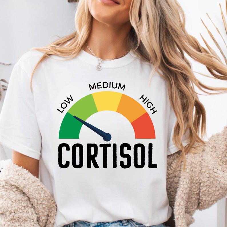 Puede incluir: Camiseta blanca con un gr&aacute;fico de un medidor de nivel de cortisol. El medidor muestra niveles de verde (bajo) a rojo (alto), con la palabra "CORTISOL" en letras negras en negrita. La camiseta la lleva una persona.