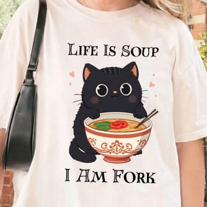 Puede incluir: Camiseta blanca con un gato negro de dibujos animados sosteniendo un cuenco de ramen. El texto "LIFE IS SOUP" está encima del gato, y "I AM FORK" debajo. El gato tiene ojos grandes y redondos y está rodeado de pequeños corazones rosas.
