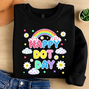 May include: Black sweatshirt with a colorful graphic that says "Happy Dot Day." The design includes a rainbow, clouds, daisies, and polka dots in various colors. The sweatshirt is made of a soft material.