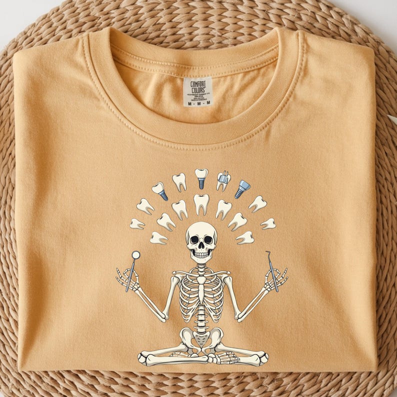 May include: Mustard-coloured t-shirt featuring a skeleton in a seated yoga pose, surrounded by teeth and dental tools. The skeleton holds a dental mirror and a probe. The shirt has a Comfort Colors tag.