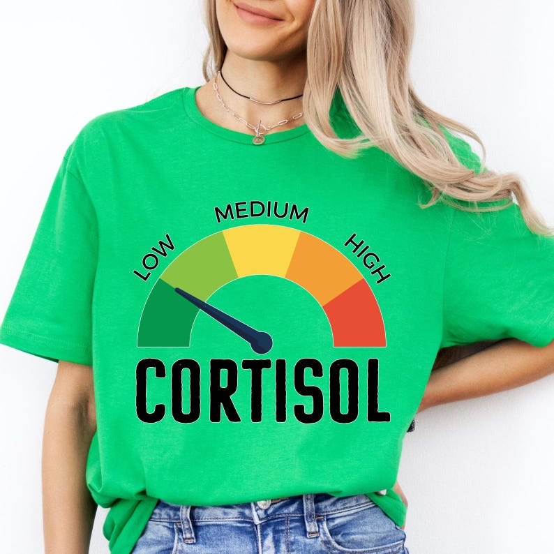 Puede incluir: Una camiseta verde con un gr&aacute;fico de un medidor de nivel de cortisol. El medidor muestra niveles de bajo a alto, con una aguja que indica el nivel medio. La palabra "CORTISOL" est&aacute; impresa en negrita debajo.