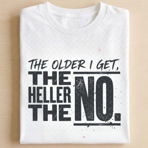 Op de afbeelding: Een wit T-shirt met korte mouwen en de zwarte tekst "THE OLDER I GET, THE HELLER THE NO." op de voorkant. Het ontwerp heeft een verweerde, getextureerde uitstraling met horizontale lijnen rond het woord "NO." en een subtiel geometrisch patroon.