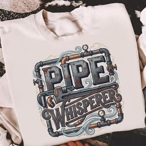 May include: A cream-coloured sweatshirt featuring the text "PIPE WHISPERER" in a steampunk-style design. The graphic includes pipes, a wrench, and gauges in grey, brown, and copper tones. The sweatshirt is made of a soft material.