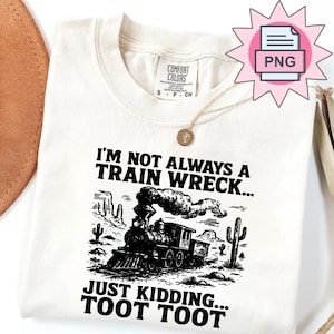 May include: Off-white t-shirt featuring a black illustration of a train in a desert scene. The text says "I'M NOT ALWAYS A TRAIN WRECK... JUST KIDDING... TOOT TOOT". A gold necklace is draped over the shirt.