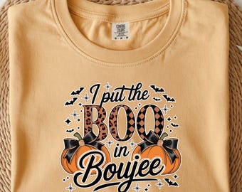 Może przedstawiać: Musztardowa koszulka z grafiką o tematyce Halloween. Projekt zawiera frazę "I put the BOO in Boujee" z akcentami nietoperzy i gwiazd oraz dwie pomarańczowe dynie z czarnymi kokardami.