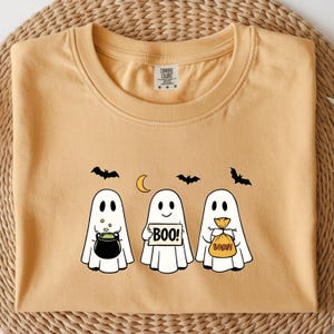 May include: Mustard-colored t-shirt featuring three cartoon ghosts with Halloween-themed accessories. One ghost holds a cauldron, another a sign that says "BOO!", and the third a bag of candy. Black bats and a crescent moon are also featured.