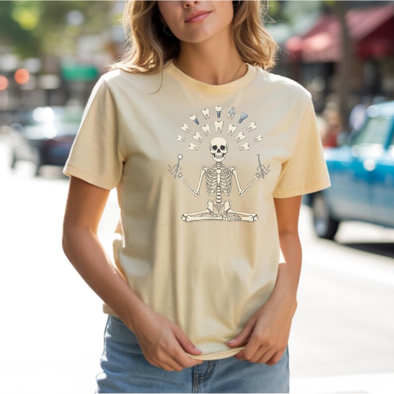 May include: A pale yellow t-shirt with a skeleton graphic in a yoga pose. The skeleton has a halo of teeth and dental tools. The design is in black and white. The shirt is made of a soft material.