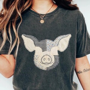 May include: A charcoal gray short-sleeve t-shirt featuring a central graphic of a pig head composed of patchwork fabric patterns, including black and white gingham and a small floral print, outlined with visible decorative stitching.