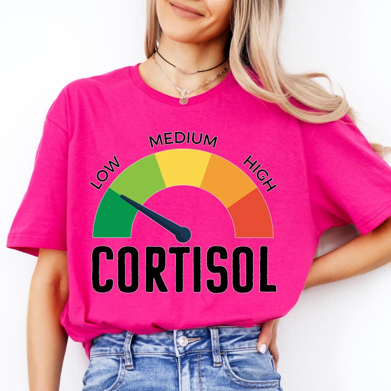 Puede incluir: Una camiseta rosa brillante con un dise&ntilde;o de medidor que muestra los niveles "BAJO", "MEDIO" y "ALTO", y la palabra "CORTISOL" en negro. La aguja del medidor apunta hacia la secci&oacute;n verde.