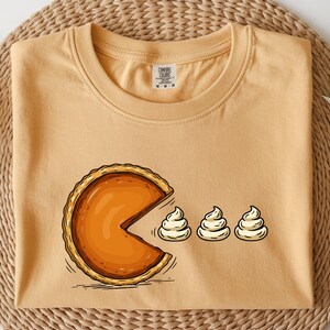 May include: Mustard-colored t-shirt featuring a cartoon pumpkin pie design. The pie is depicted with a Pac-Man-like bite taken out of it, with three dollops of whipped cream. The shirt has a crew neck and short sleeves.