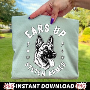 May include: A pale green t-shirt with a German Shepherd dog illustration and the text "EARS UP SYSTEM ARMED". The design includes lightning bolts and star emblems. The image is a PNG instant download.