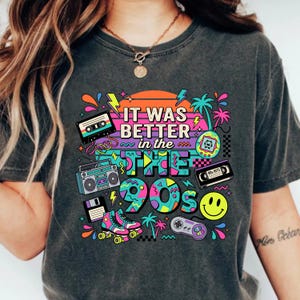 May include: A charcoal gray t-shirt featuring a colorful 1990s-themed graphic print. The design includes a cassette tape, boombox, floppy disk, roller skates, handheld game, VHS tape, smiley face, and game controller with the text It Was Better in the 90s.