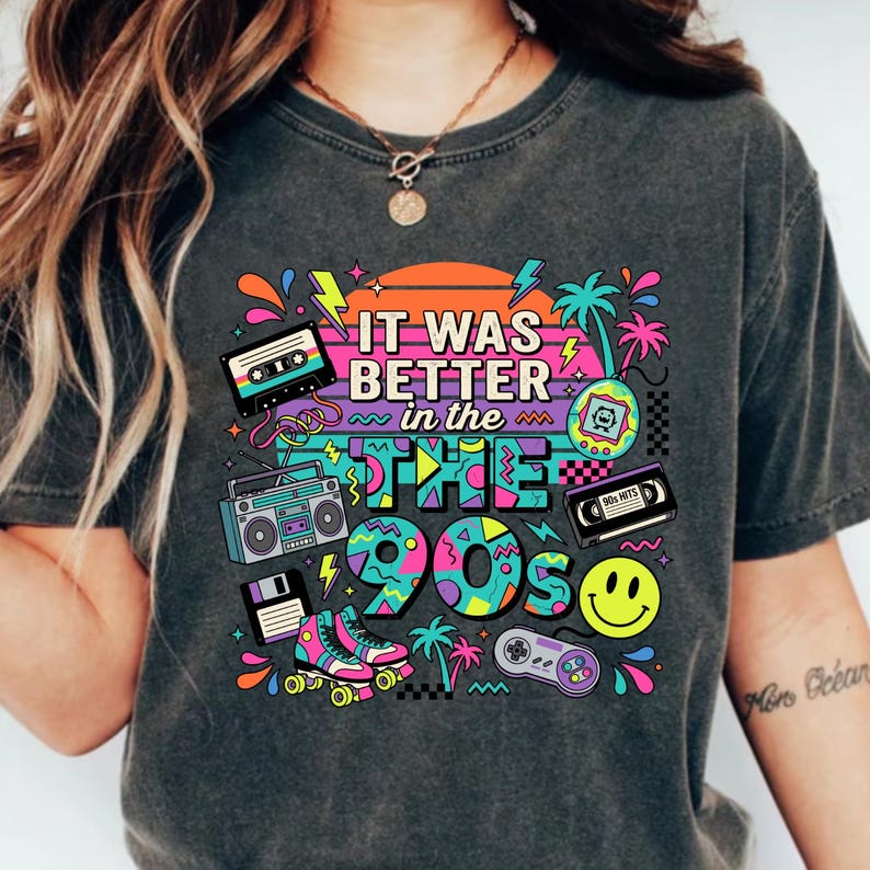May include: A charcoal gray t-shirt featuring a colorful 1990s-themed graphic print. The design includes a cassette tape, boombox, floppy disk, roller skates, handheld game, VHS tape, smiley face, and game controller with the text It Was Better in the 90s.