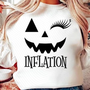Op de afbeelding: Witte sweatshirt met een zwart pompoengezicht ontwerp. Het ontwerp omvat een driehoekige neus, een knipogend oog met lange wimpers en het woord "INFLATION". Een dollarteken staat boven het knipogende oog.