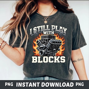 May include: Dark gray t-shirt with the text "I STILL PLAY WITH BLOCKS" in white. The graphic features a detailed engine design with flames. The shirt is a casual wear item.