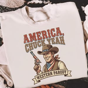 May include: Cream-colored sweatshirt featuring a graphic of a smiling cowboy holding a gun. The text "AMERICA, CHUCK YEAH" is above the cowboy, and "WESTERN PARODY" is below. The cowboy wears a hat, vest, and a bandana with a USA flag design.