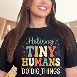 May include: Black t-shirt with the text "Helping TINY HUMANS DO BIG THINGS" in a retro font. The design includes stars, pencils, an apple, a book, and flowers. A fun and educational themed shirt.