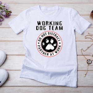 May include: White t-shirt with the text "WORKING DOG TEAM" above a circular design. The design features a paw print and the words "DO NOT DISTRACT PARTNER AT WORK" in a red circle.
