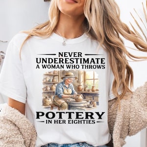 Puede incluir: Una camiseta blanca con una ilustración gráfica de una persona mayor con sombrero y delantal, trabajando en un torno de alfarero en un taller. El texto dice: NEVER UNDERESTIMATE A WOMAN WHO THROWS POTTERY IN HER EIGHTIES.