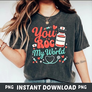 Może przedstawiać: Ciemnoszara koszulka z kolorową grafiką z napisem "You Roc My World". Projekt zawiera strzykawkę, stetoskop, serca i fiolkę z napisem "Rocuroium". Koszulka ma wygląd vintage.