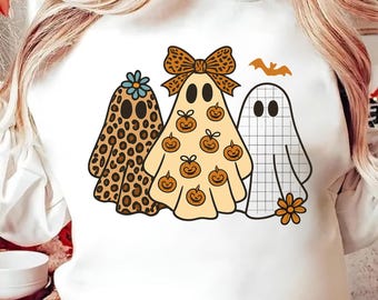 May include: White sweatshirt featuring three cartoon ghosts. One ghost has a leopard print design, another has pumpkin details, and the third has a grid pattern. A bat and flower complete the Halloween-themed design.