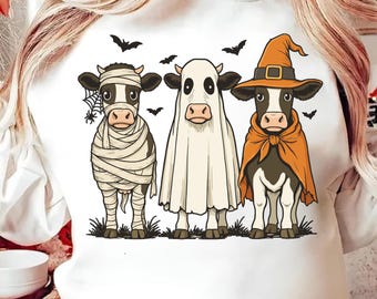 May include: White sweatshirt featuring a Halloween-themed graphic of three cows dressed in costumes: a mummy, a ghost, and a witch. Black bats and a spiderweb are also included in the design. The cows are standing on green grass.