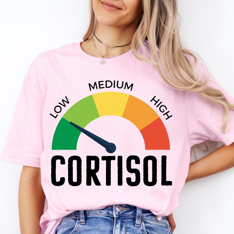 Puede incluir: Camiseta rosa claro con un dise&ntilde;o de medidor y la palabra "CORTISOL" en negro. La aguja del medidor apunta a la secci&oacute;n verde, etiquetada como "LOW". La parte superior del medidor est&aacute; etiquetada como "MEDIUM" y "HIGH".