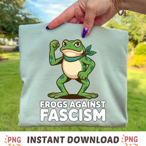 May include: A pale blue t-shirt with a cartoon frog design and the words "FROGS AGAINST FASCISM". The frog is green with darker green spots and a cream-colored belly. The shirt is being held up by a person.