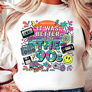 May include: A white sweatshirt featuring a colorful graphic print with the text IT WAS BETTER in the 90s. The design includes a cassette tape, boombox, floppy disk, roller skates, handheld game, VHS tape, game controller, and a yellow smiley face.