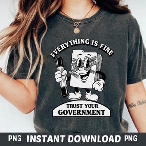 Peut inclure: T-shirt gris foncé avec un personnage de dossier de dessin animé tenant un stylo. Le texte "EVERYTHING IS FINE" est au-dessus et "TRUST YOUR GOVERNMENT" en dessous. Le dossier contient des étiquettes "TOP SECRET" et "CLASSIFIED".