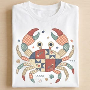 May include: White t-shirt featuring a colorful crab design. The crab is made of patchwork patterns in red, blue, and tan, with star and seashell accents. A whimsical, nautical-themed graphic tee.