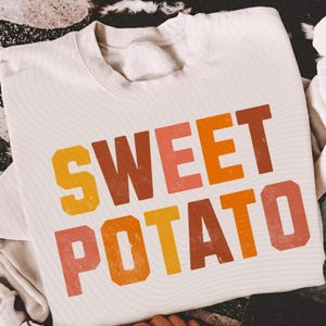 Op de afbeelding: Crèmekleurige sweatshirt met de tekst "SWEET POTATO" in grote letters in retrostijl. De tekst gebruikt een verloop van oranje, gele en roze tinten. De sweatshirt heeft een ronde hals en lange mouwen.