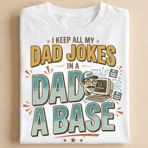 May include: A white t-shirt featuring a graphic with the text I KEEP ALL MY DAD JOKES IN A DAD A BASE. The design includes an illustration of a person wearing a cap, a vintage computer monitor labeled DATABASE, and two floppy disks labeled JOKES and FUN.