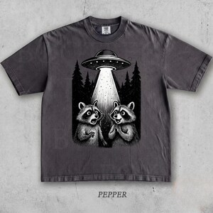 May include: Dark grey t-shirt with a black and white graphic of two raccoons gazing at a UFO. The UFO emits a bright light beam. The word "PEPPER" is printed at the bottom of the shirt.