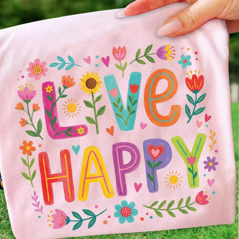 K&ouml;nnte beinhalten: Hellrosa T-Shirt mit den Worten "LIVE HAPPY" in bunten, floral verzierten Buchstaben. Das Design umfasst Blumen, Herzen und Sonnen und schafft eine fr&ouml;hliche und lebendige &Auml;sthetik.