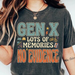 May include: A charcoal gray short-sleeve t-shirt featuring a retro-style graphic with the text "GEN X LOTS OF MEMORIES NO EVIDENCE" in blue and orange. The design includes small icons of a pixelated alien, a bicycle, and a cassette tape.