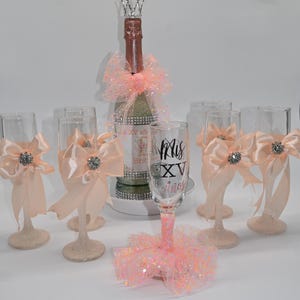 Peut inclure: Ensemble de flûtes à champagne décorées de paillettes roses et de nœuds. Une bouteille de champagne est décorée de paillettes roses et d'une couronne. Une flûte porte l'inscription "Mis XV Anos".